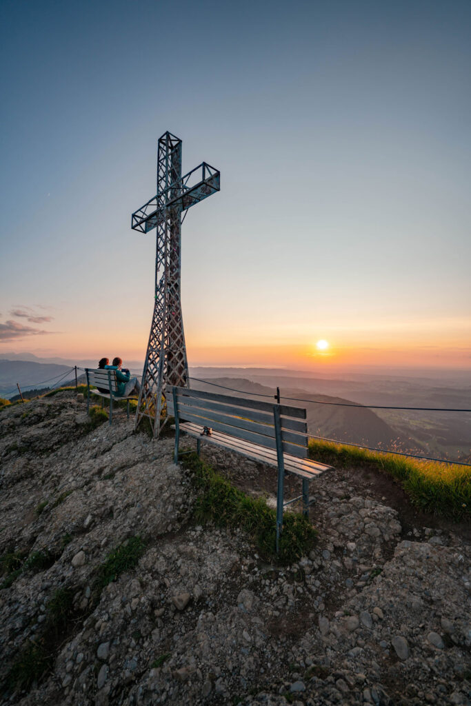 Zwei Wanderer sitzen auf einer Holzbank am Hochgrat und genießen die weite Panorama-Aussicht auf die Allgäuer Alpen.