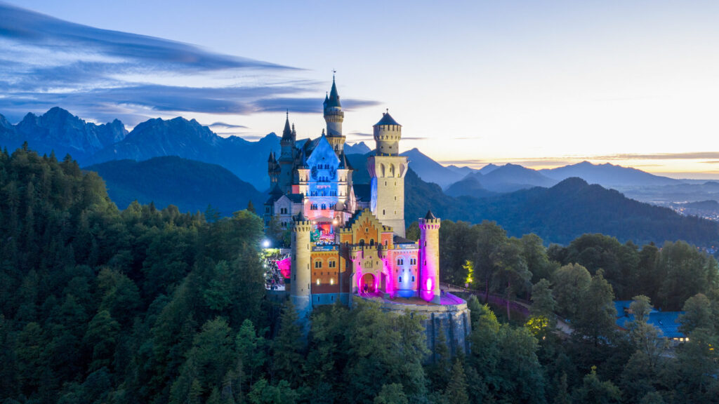 Luftaufnahme des farbig beleuchteten Schlosses Neuschwanstein bei Nacht während einer Abendveranstaltung.