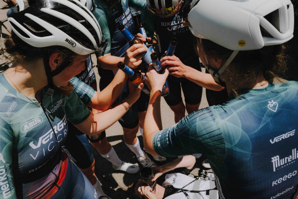 Eine Gruppe von Radsportlern im Teamtrikot stößt nach dem Radrennen kreisförmig mit Flaschen Erdinger Alkoholfrei von oben fotografiert an.