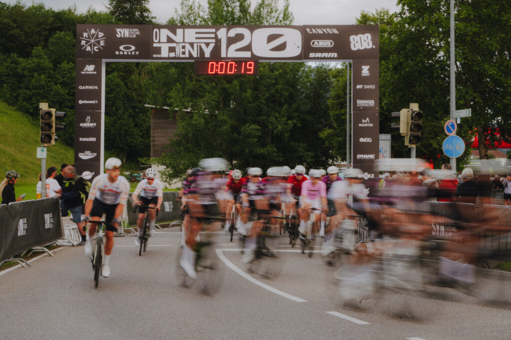 Dynamischer Massenstart des Radrace One-Twenty Rennen mit verschwommener Bewegungsunschärfe unter dem Sponsoren-Zielbogen.