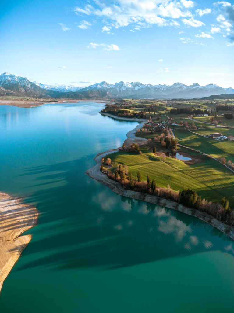 Weitwinkel-Luftaufnahme des türkisfarbenen Forggensees im Frühling mit Blick auf die schneebedeckte Tannheimer und Allgäuer Alpenkette.