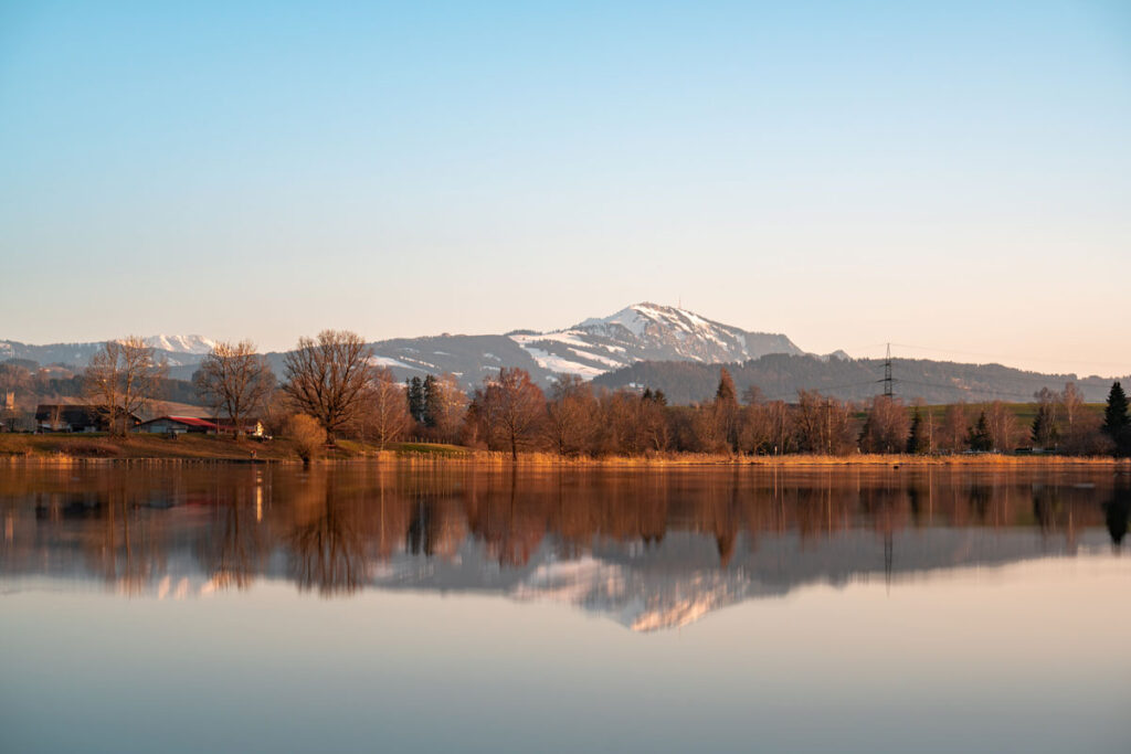 Eine perfekte Spiegelung der schneebedeckten Berge und Bäume auf der glatten Wasseroberfläche des Öschlesees bei Sulzberg in der Abendsonne.