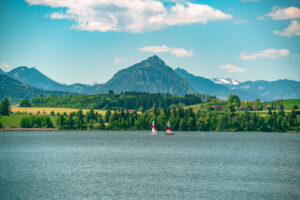 Zwei Segelboote gleiten über den Hopfensee bei Füssen vor der Kulisse der Allgäuer Voralpen unter blauem Himmel.