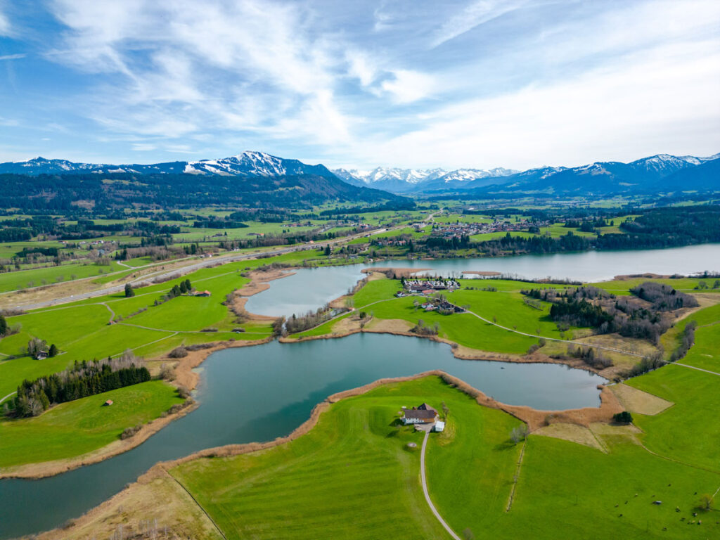 Vogelperspektive auf den Niedersonthofener See umgeben von sattgrünen Wiesen und mit Blick auf die schneebedeckten Allgäuer Alpen.