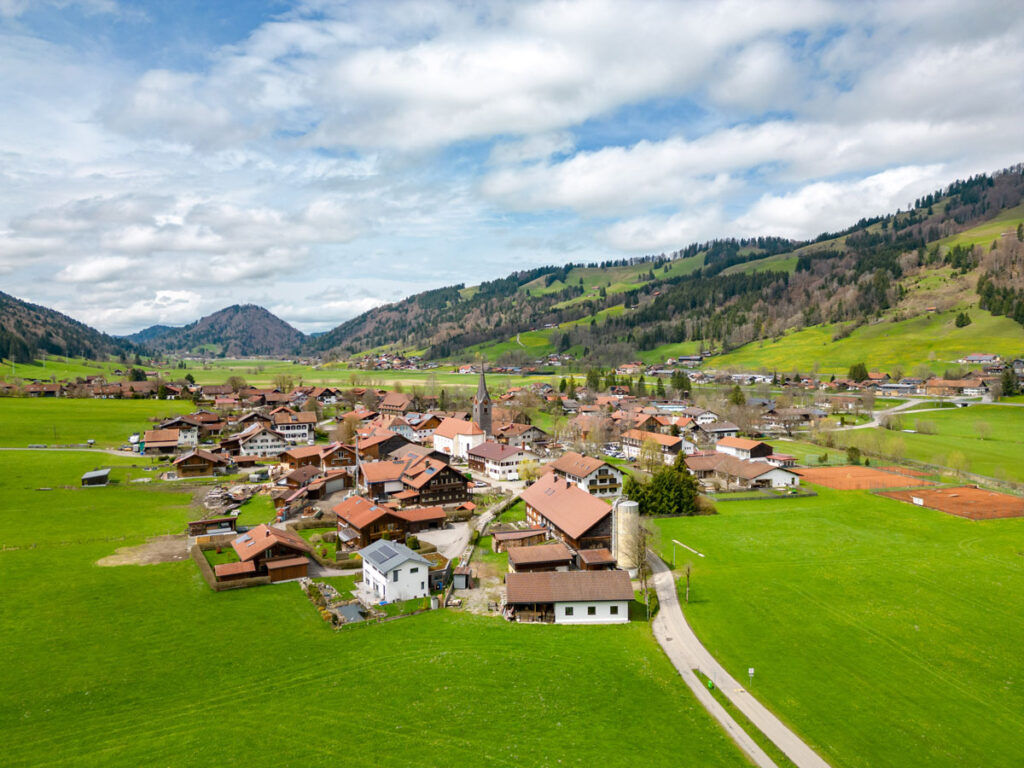 Vogelperspektive aus der Drohne auf das idyllische Dorf Thalkirchdorf bei Oberstaufen inmitten grüner Frühlingswiesen.