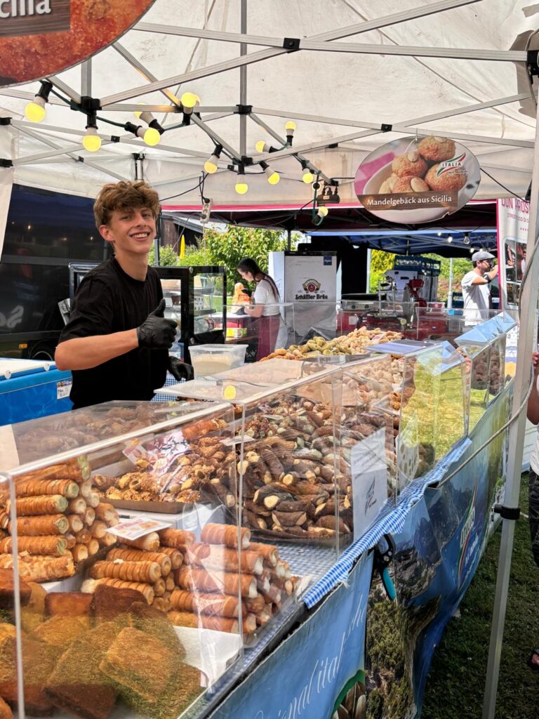 Verkäufer lächelt hinter einem Marktstand voller sizilianischer Cannoli und Mandelgebäck auf einem Streetfood-Markt im Oberstaufen