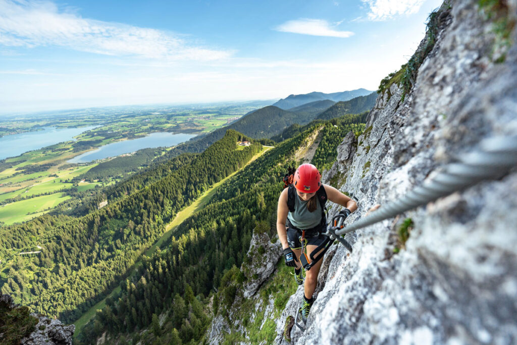 Eine Kletterin am Tegelberg-Klettersteig mit spektakulärem Tiefblick auf die Allgäuer Seenlandschaft.