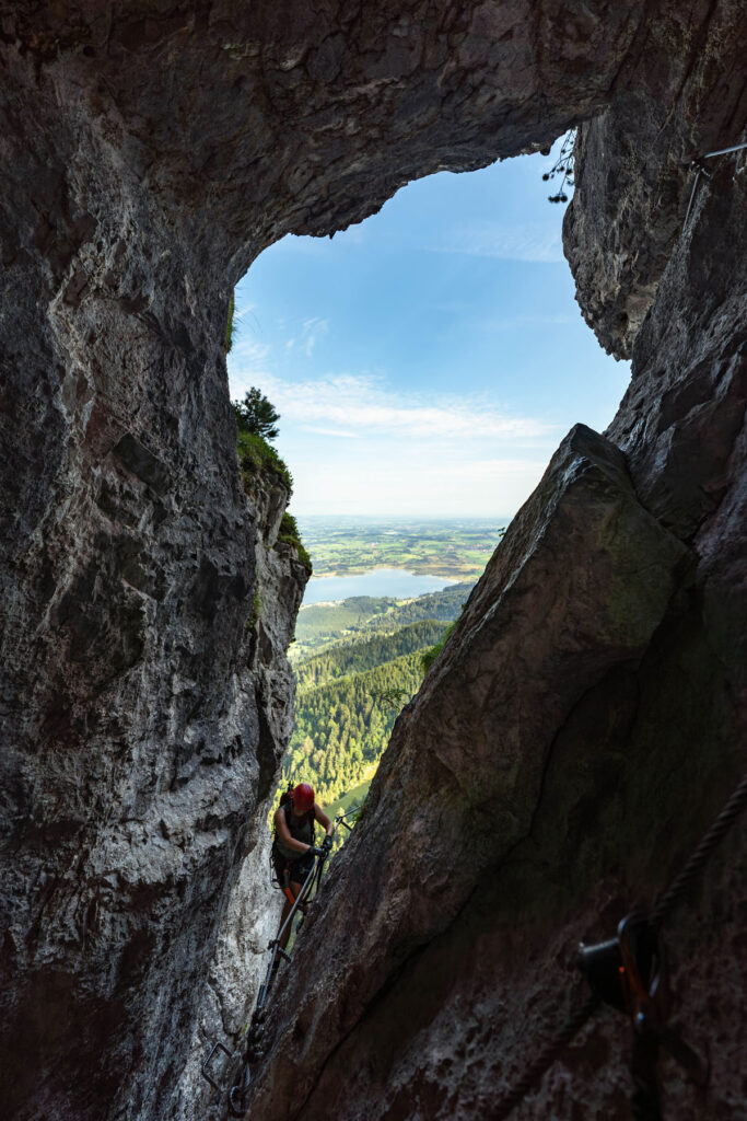 Blick durch ein imposantes Felsfenster am Tegelberg-Klettersteig auf das grüne Allgäuer Voralpenland.